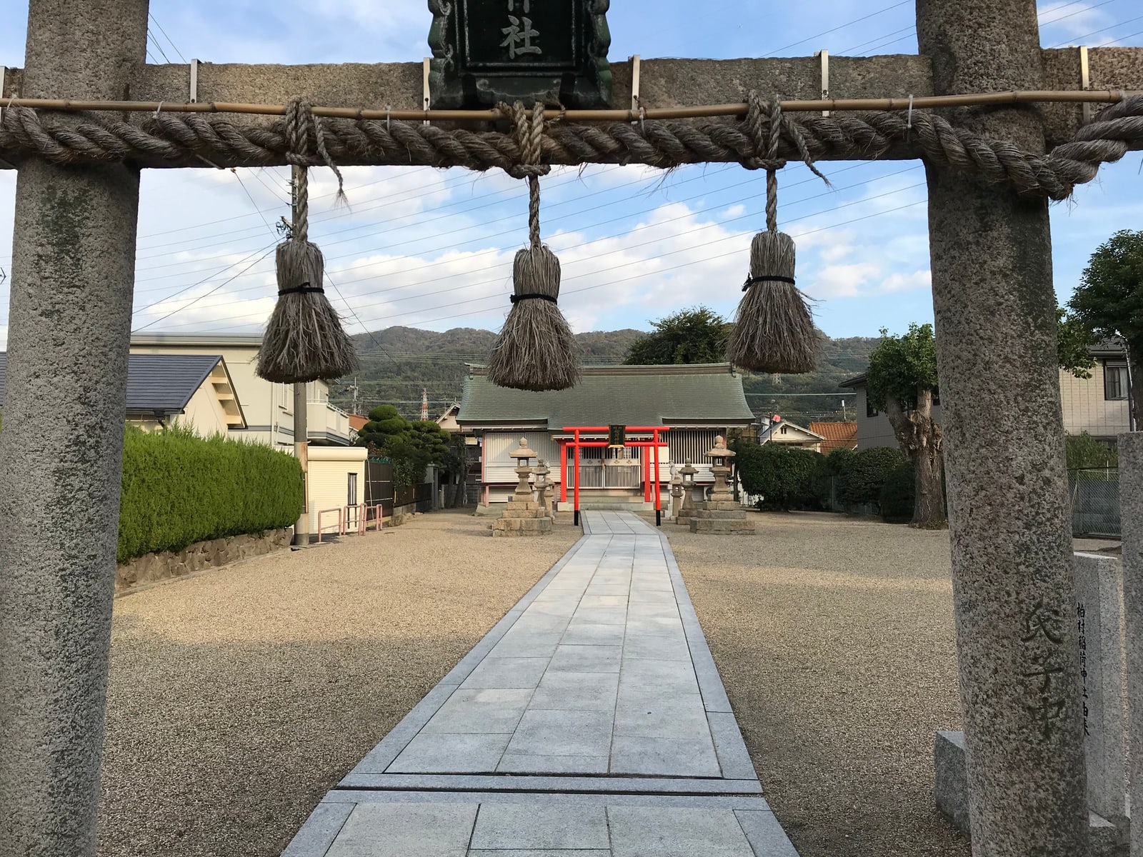 A small neighborhood Shinto shrine with a torii gate and paved approach path, houses visible beyond the grounds.