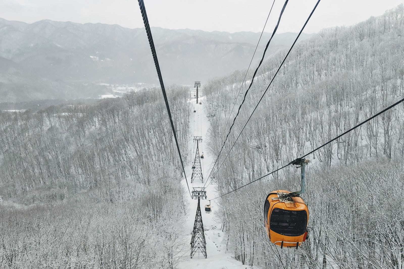 Looking down a gondola line from a cabin, cables converging to lattice towers over a cleared corridor through snow-frosted forest, a snow-covered valley and village below.