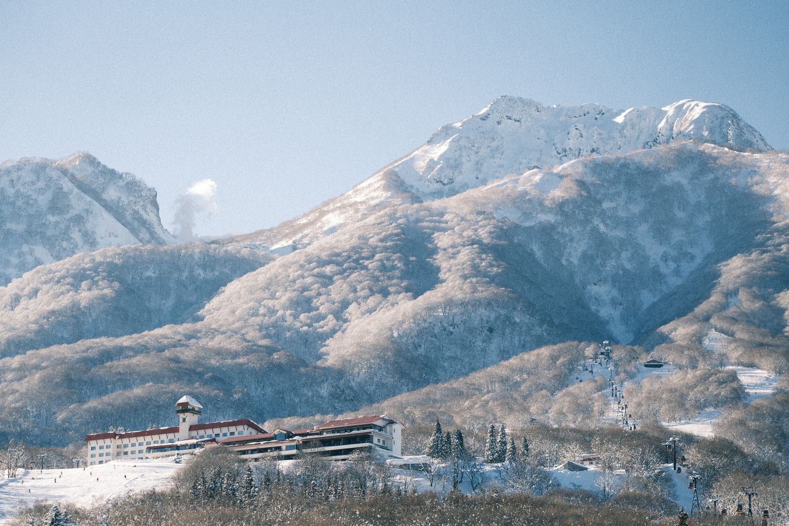 A large white resort hotel with red roofs and a clock tower below snow-covered forested slopes, a lift line and ski runs to the right, a craggy peak behind.