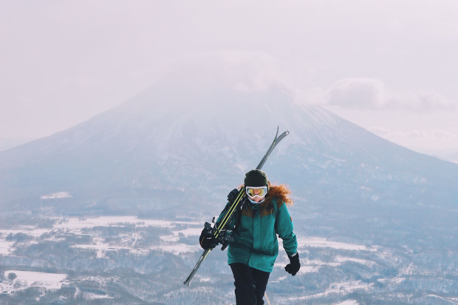 One skier in a teal jacket walking uphill with skis over the shoulder, a snow-covered valley floor and a large conical mountain behind under hazy cloud.