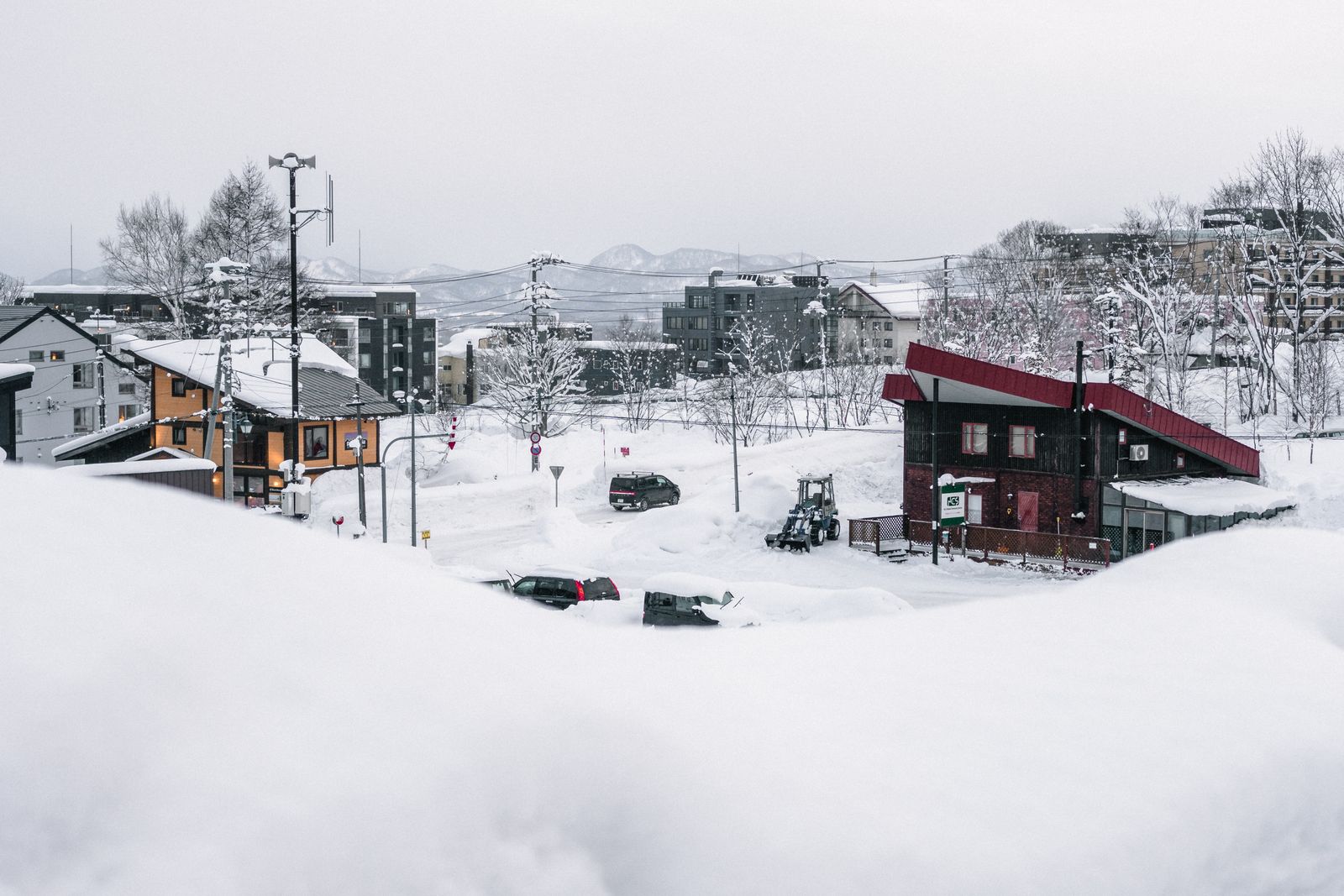 A snowed-in resort town under a white sky: deep snowbanks, two cars buried to the windows, a wheel loader, timber houses and condominium blocks behind.
