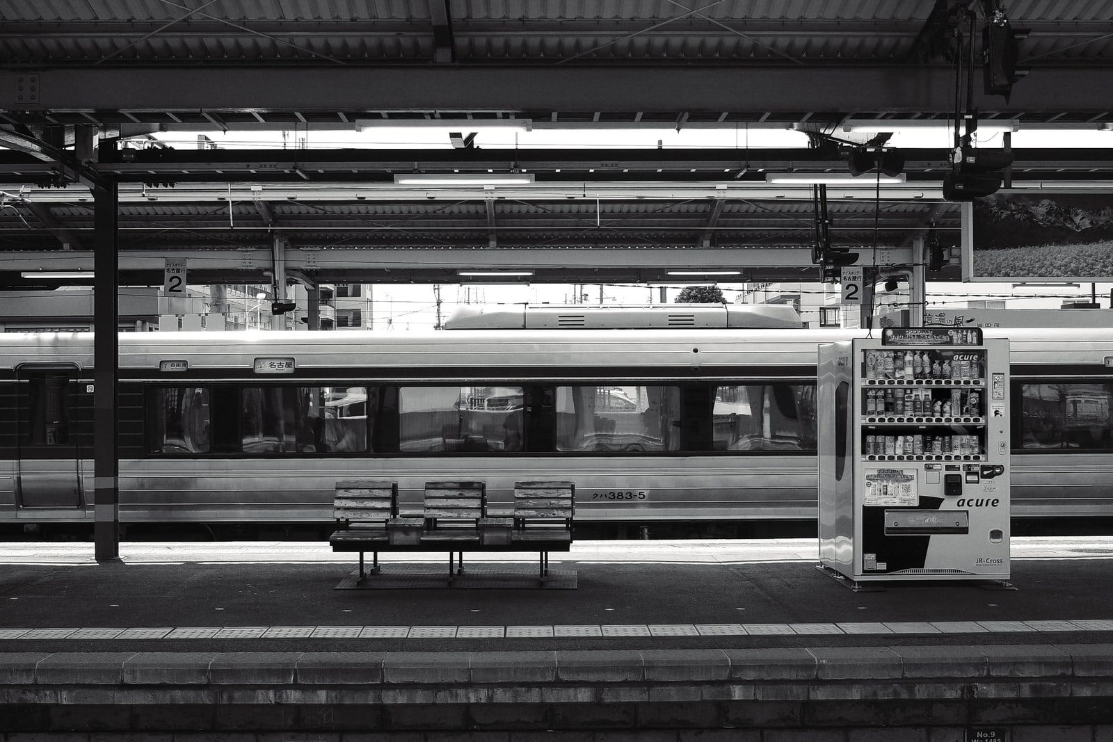 A black-and-white view of a train standing at a platform in Nagano, a bench and a vending machine in front.