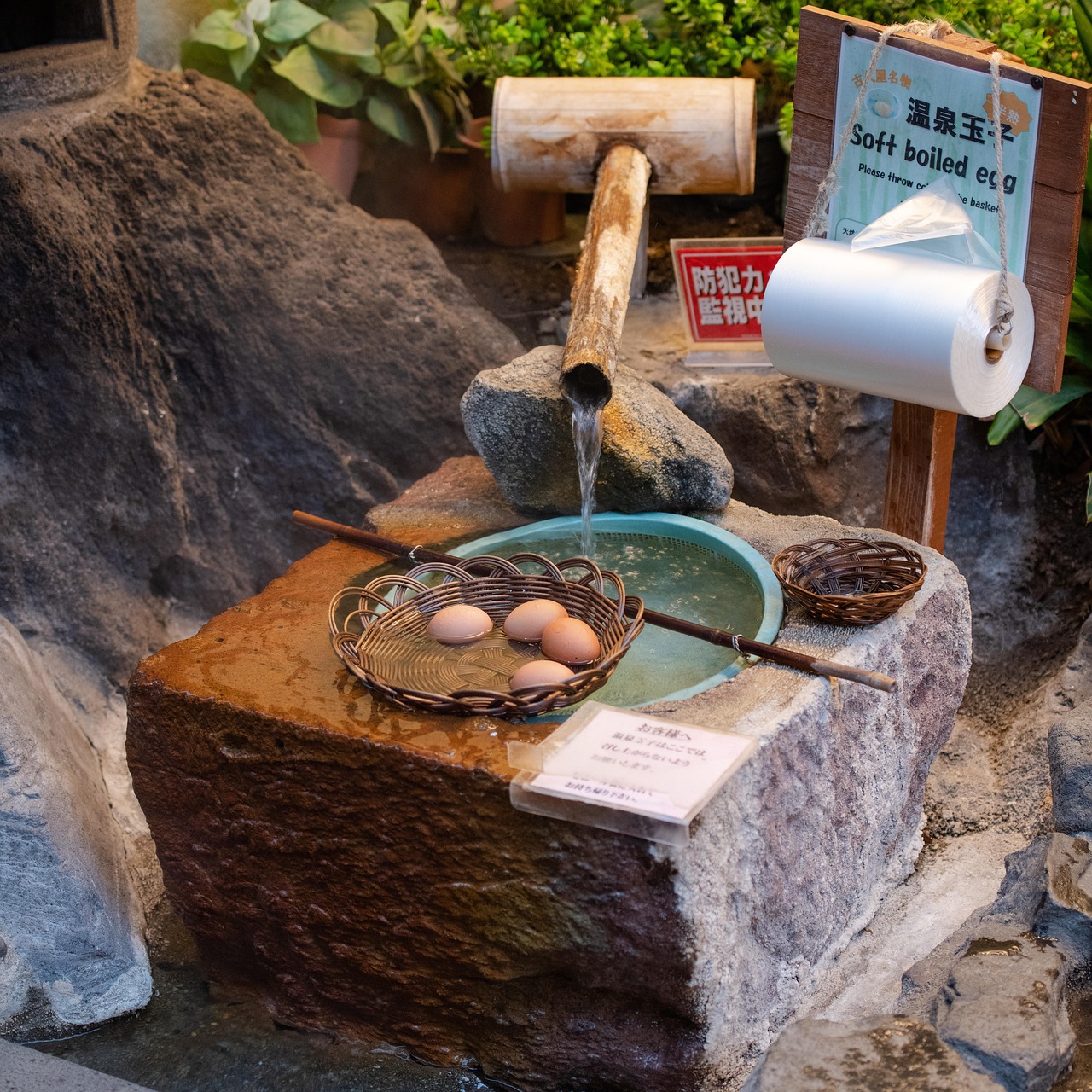 A basket of eggs cooking in a stone basin of hot spring water fed by a bamboo spout, a sign for soft-boiled eggs above.