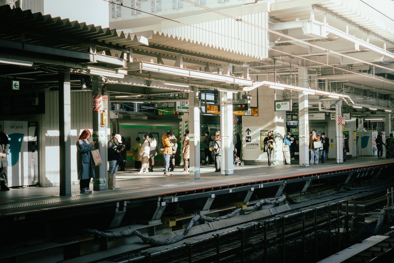 Passengers standing along a covered train platform in daylight, seen from across the tracks.