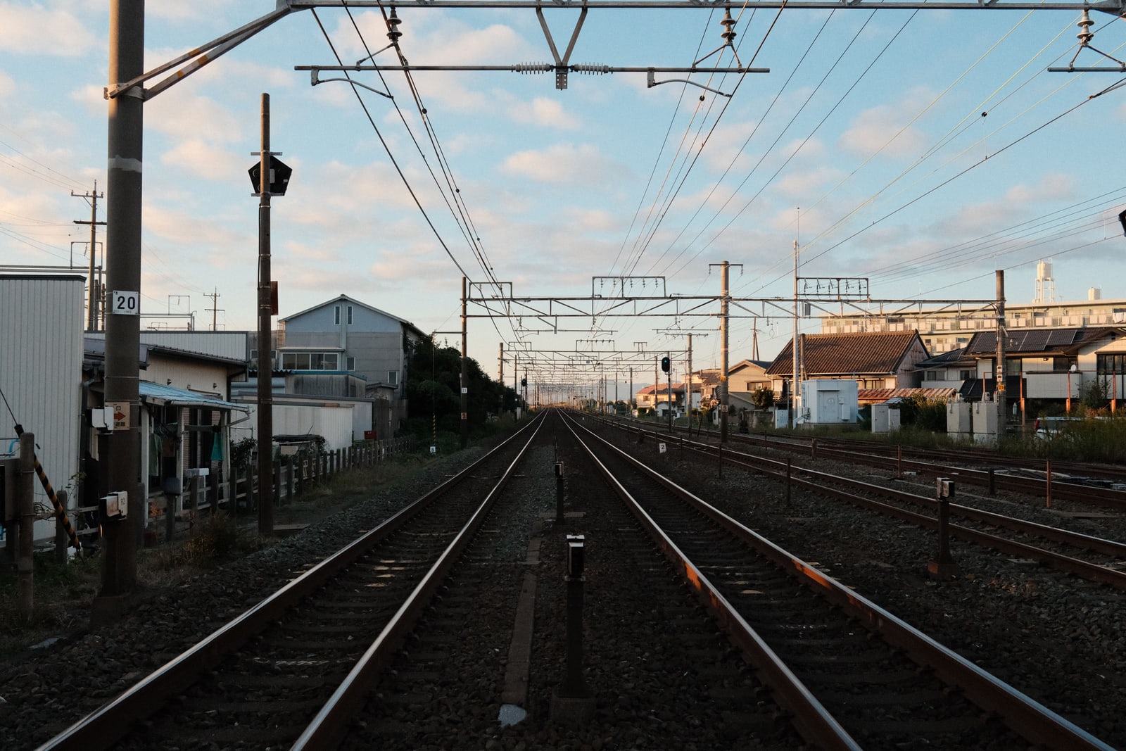 A double railway track running straight between low-rise houses under overhead wires at dusk.