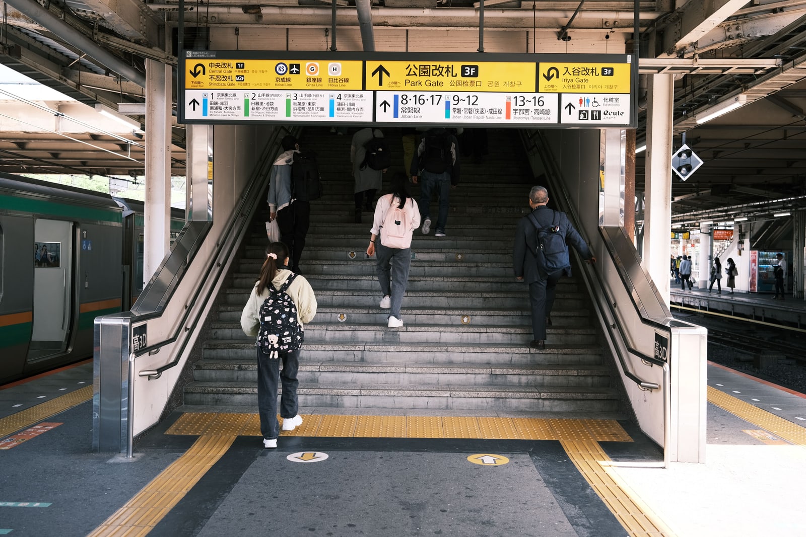 People climbing a station staircase toward yellow exit signs, seen from behind.
