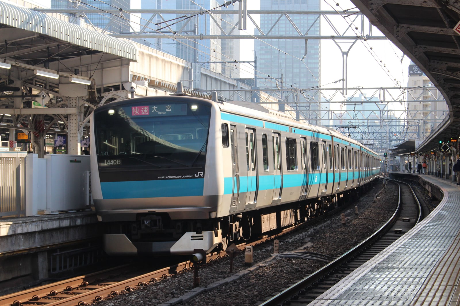 A sky-blue striped JR commuter train curving into a station platform with office towers behind it.