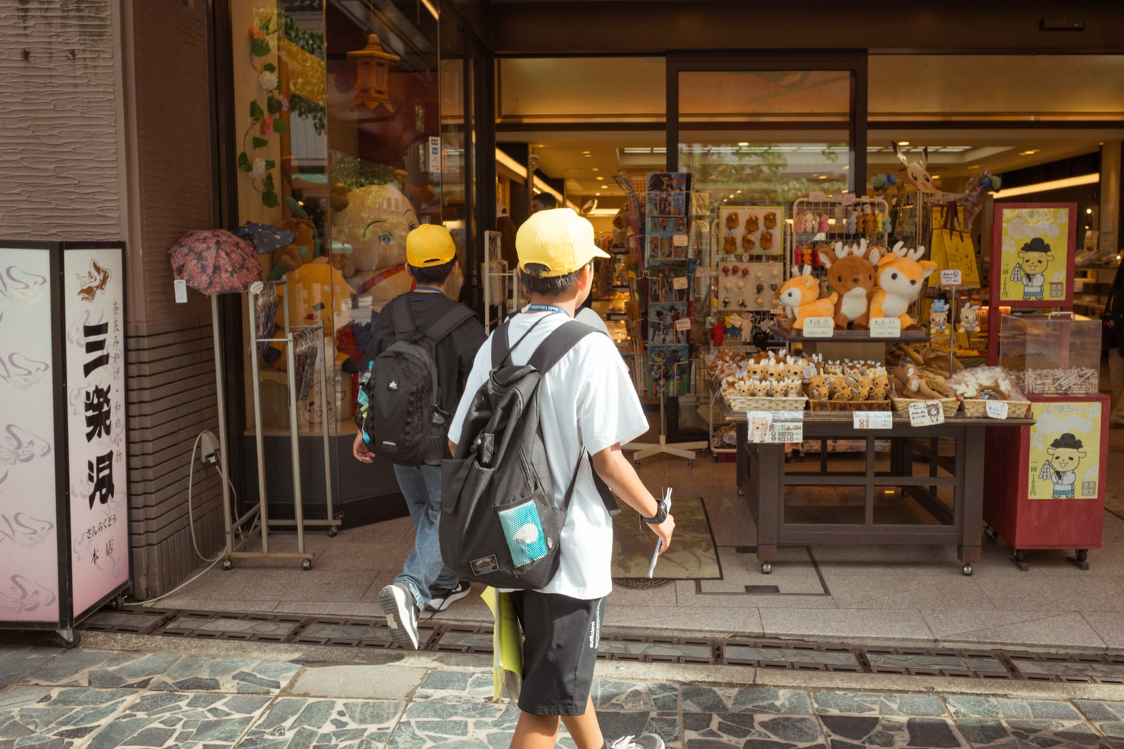 Two boys in yellow caps and backpacks, seen from behind, at the open front of a toy and souvenir shop in Japan.