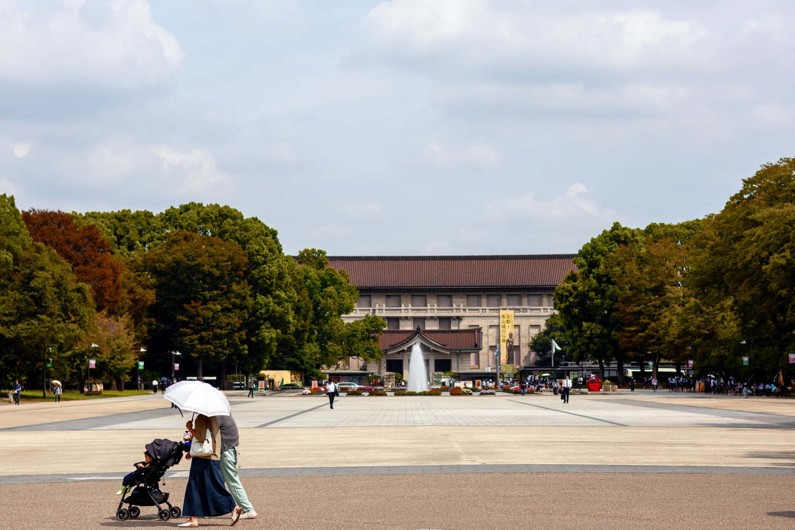 A woman with an umbrella pushing a stroller across the open plaza of Ueno Park, with the Tokyo National Museum behind.