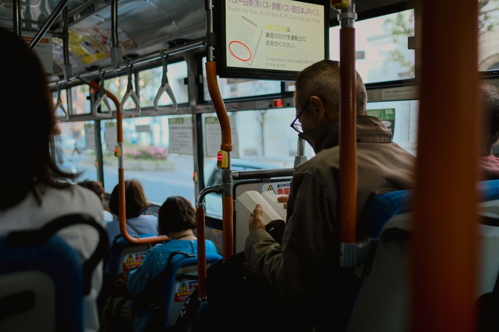 Inside a Kyoto City Bus, orange grab poles and blue seats, an overhead screen advertising the one-day bus pass, passengers seen from behind.