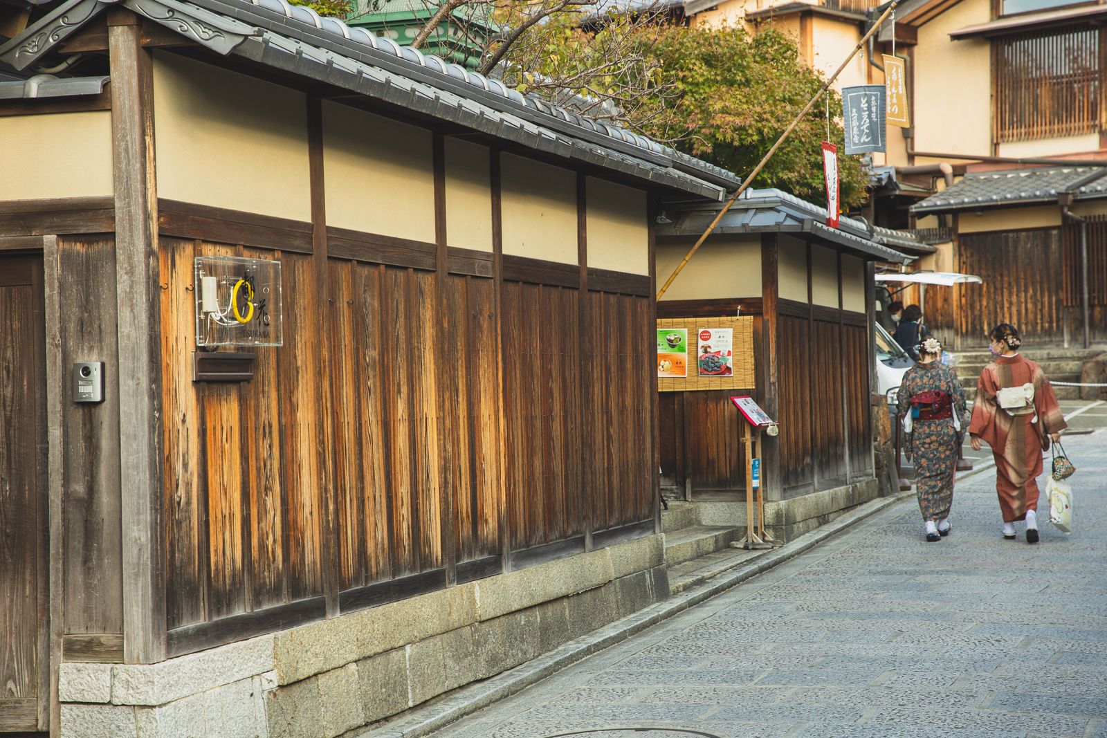 A stone-paved lane in Kyoto's preserved Higashiyama district, wooden lattice walls and tiled eaves on both sides, two women in kimono walking away from the camera.