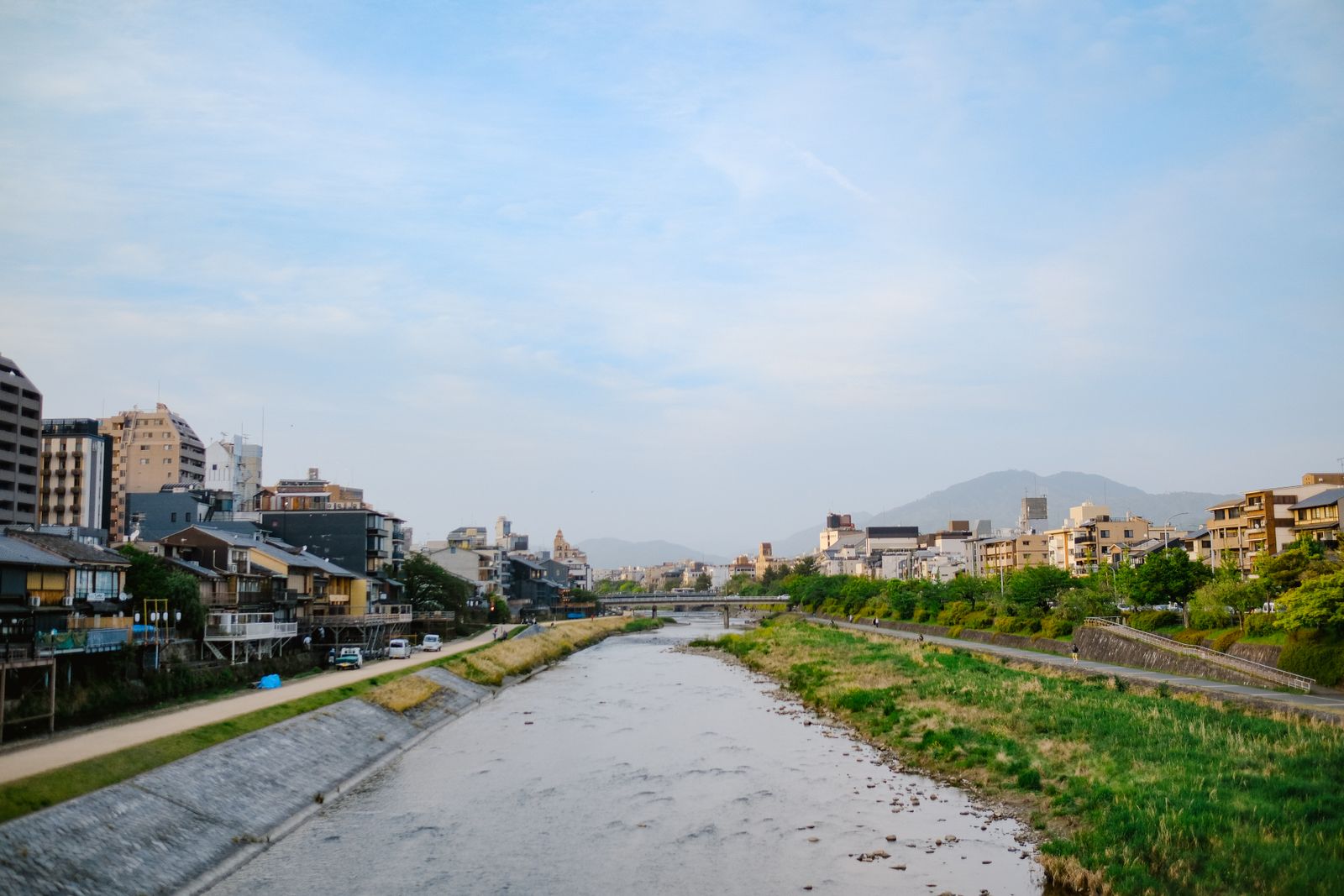 The Kamo river seen from a bridge, gravel bed and embankment paths on both banks, restaurant terraces and mid-rise hotels along the west bank and the Higashiyama hills behind.