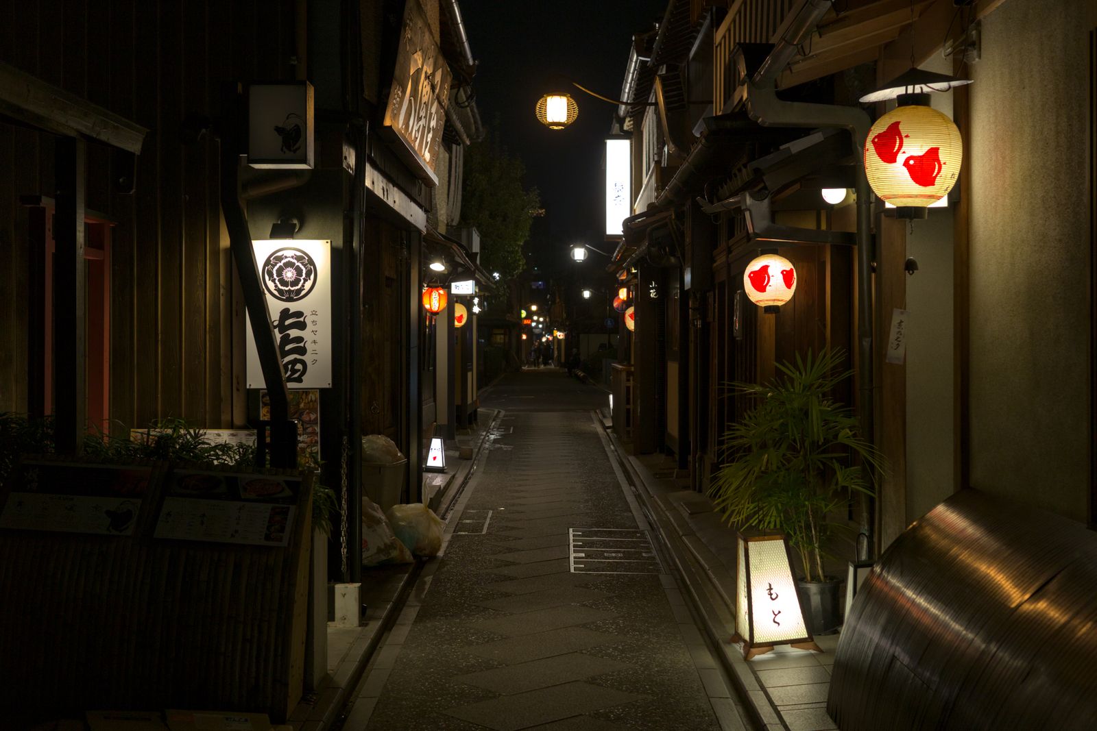 A narrow stone-paved alley at night between wooden restaurant fronts, paper lanterns with a plover emblem lighting the way.