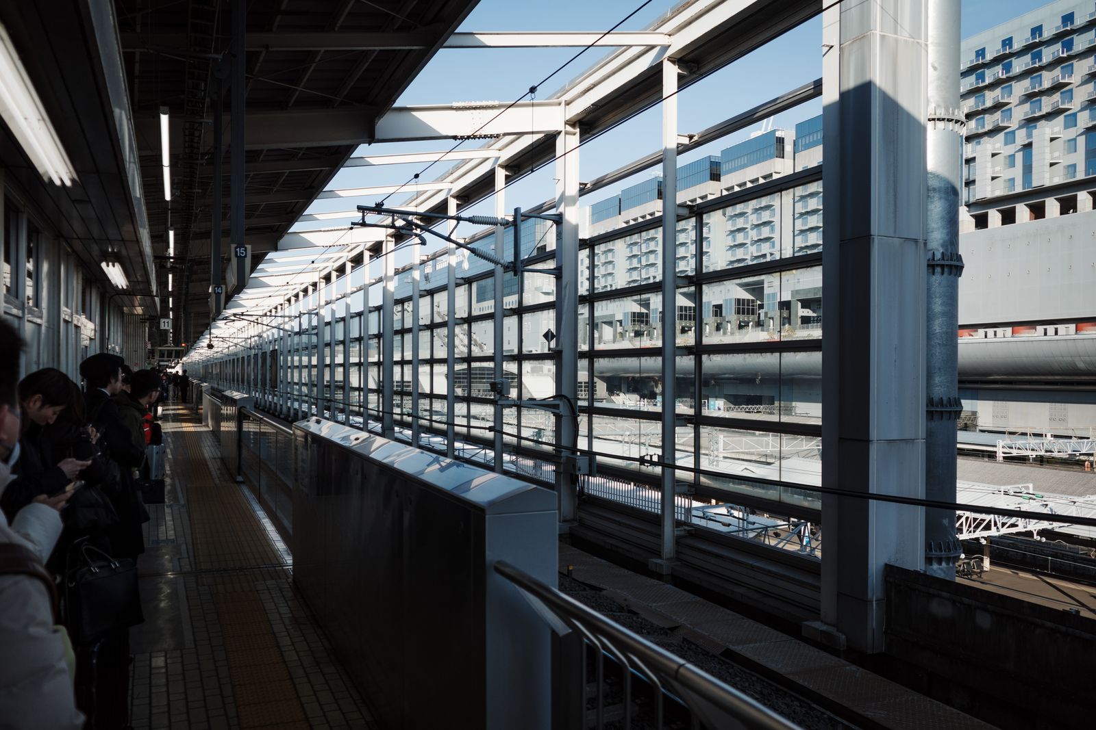 An elevated Shinkansen platform at Kyoto Station with full-height platform doors, car-stop markers hanging from the canopy, and the glass station building and hotel tower rising behind.