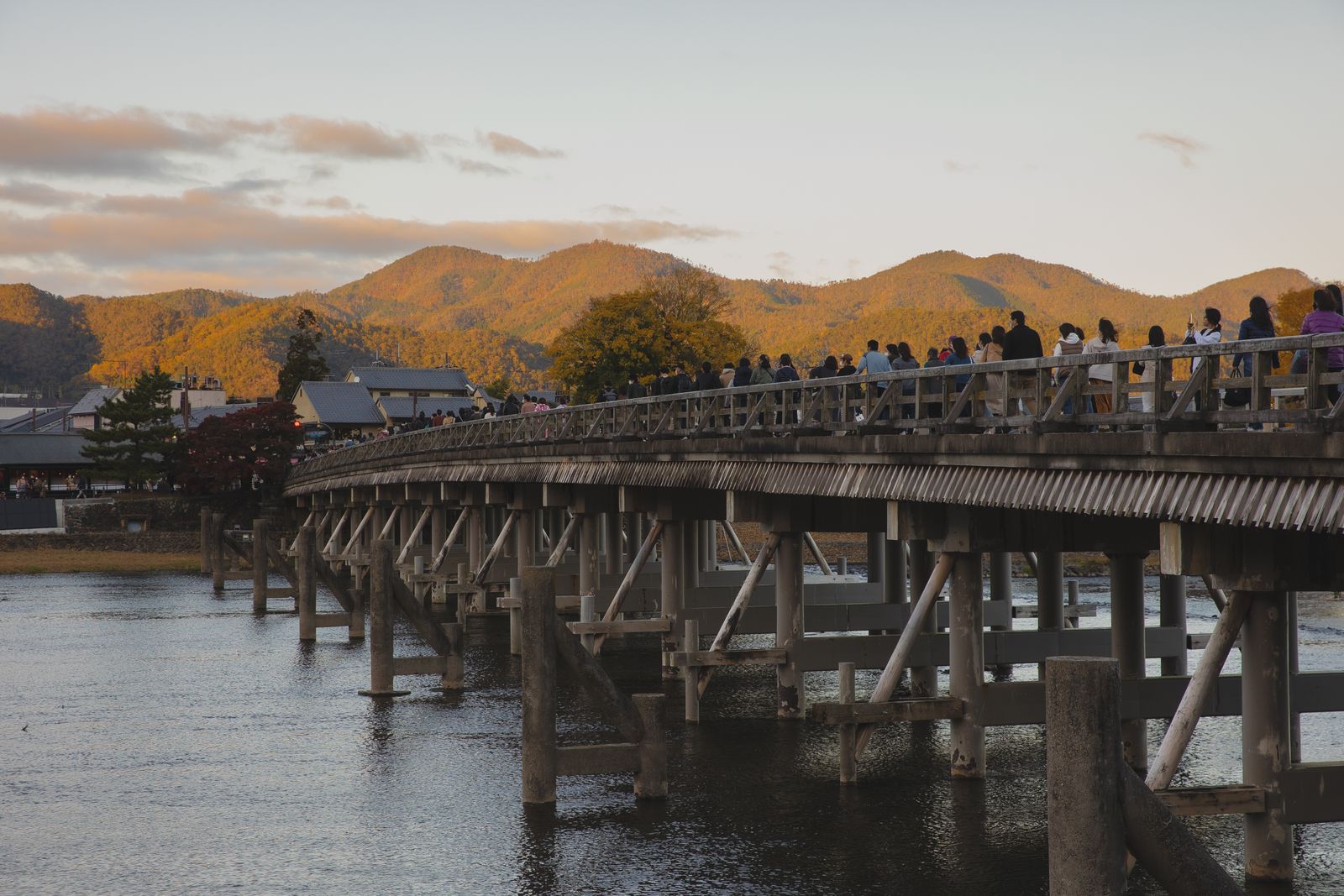 The long timber-railed Togetsukyo bridge over the Katsura river at sundown, a crowd of walkers on it and the Arashiyama hills in autumn color behind.