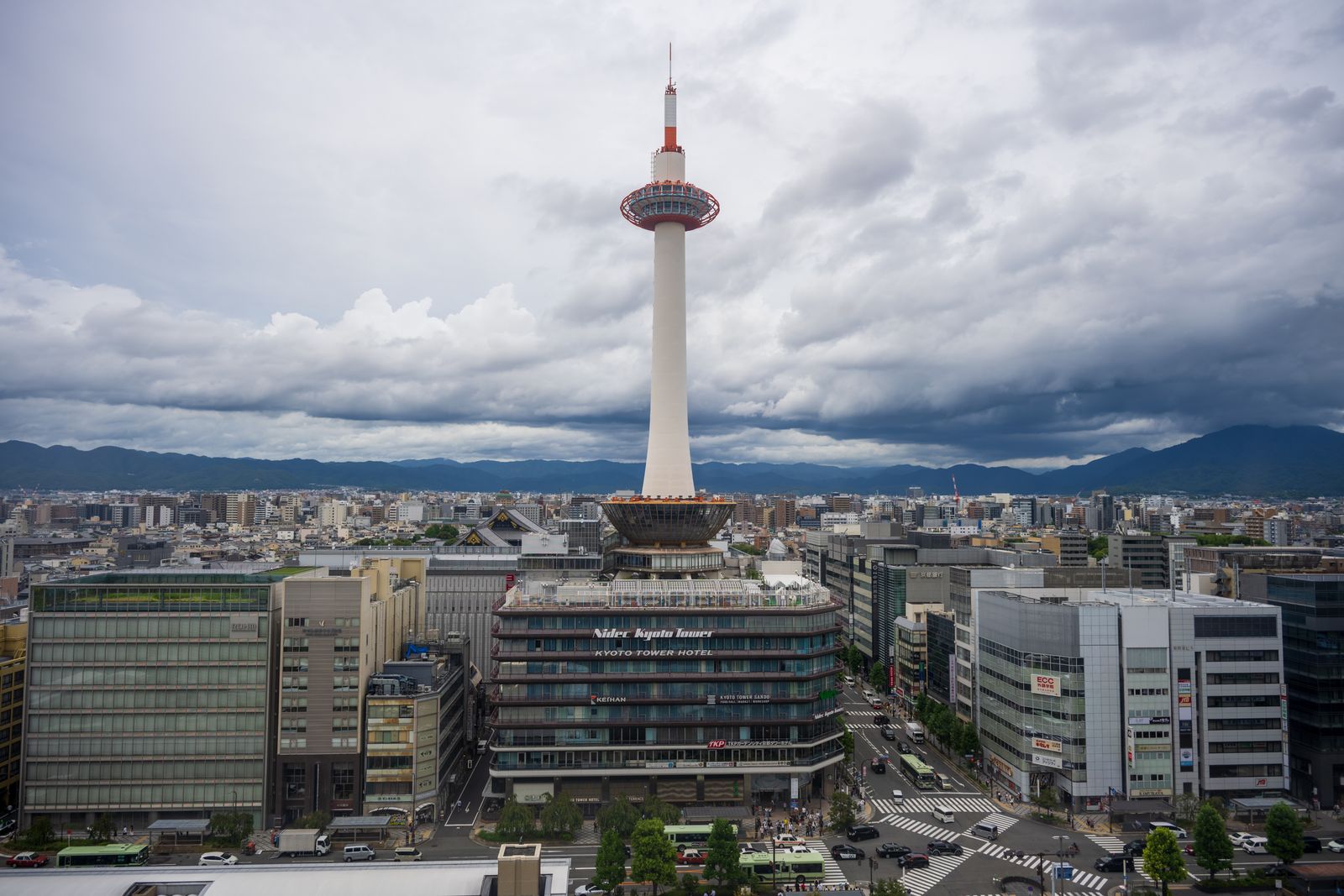 Kyoto Tower on its hotel base seen from above the station forecourt, green city buses in the street below and the northern hills under cloud.