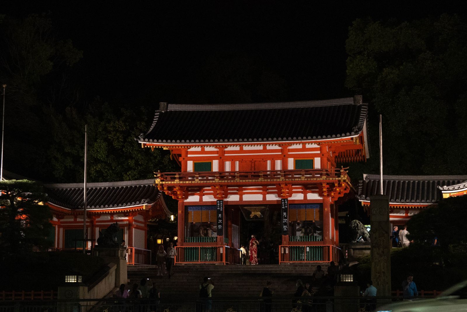 The vermilion two-story West Gate of Yasaka Shrine lit at night, banners in the gateway and a few visitors on the steps.