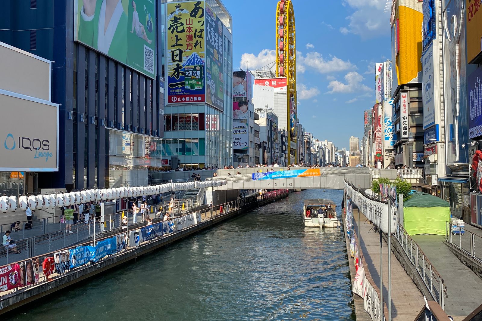 The Dotonbori canal in daylight from upstream of Ebisubashi, the bridge nameplate legible, lantern rails along both riverwalks and a small tour boat mid-channel.