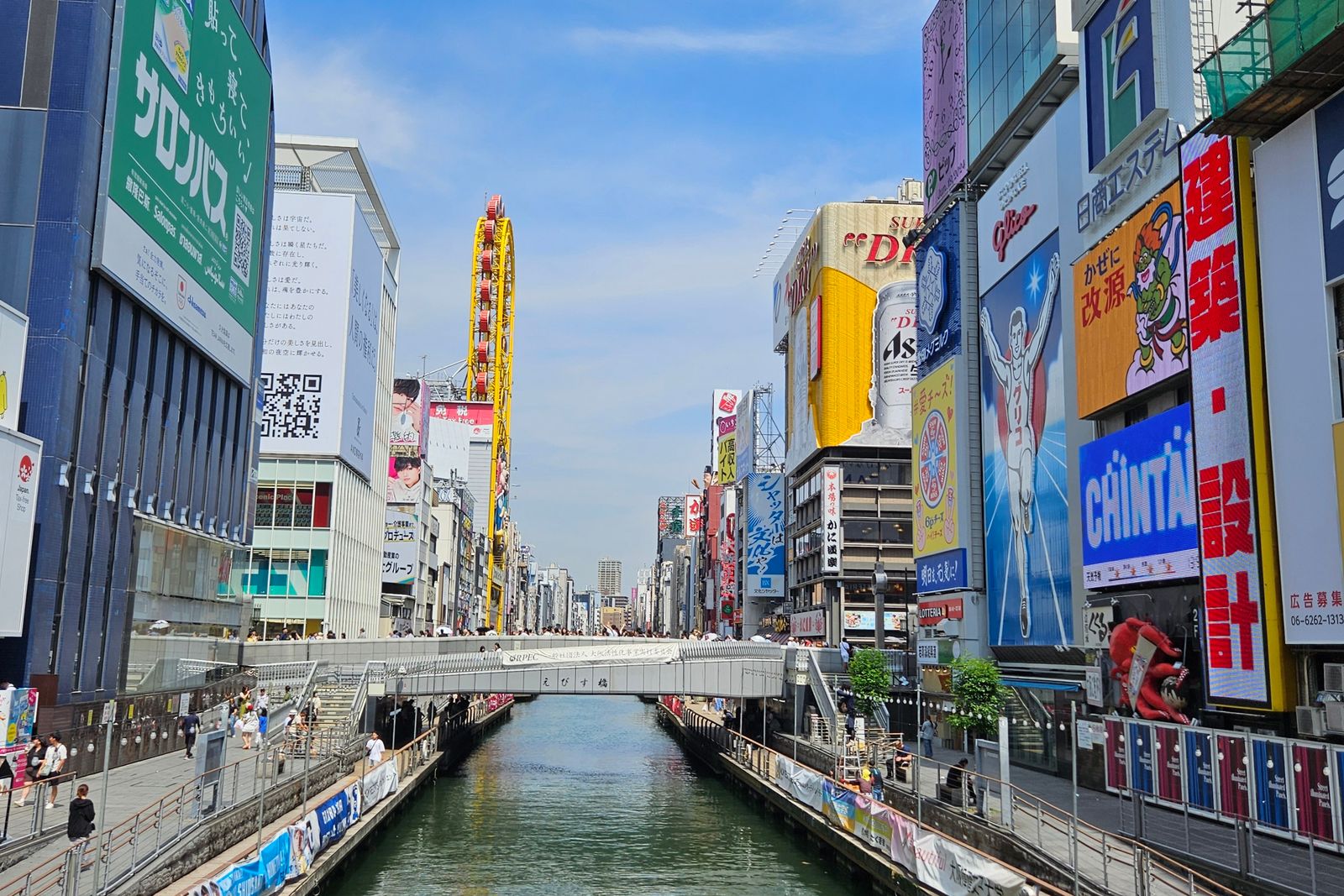 The Dotonbori canal in daylight, the Ebisubashi nameplate on the bridge parapet, the Glico running man billboard on the right bank and the Ebisu Tower ferris wheel behind.