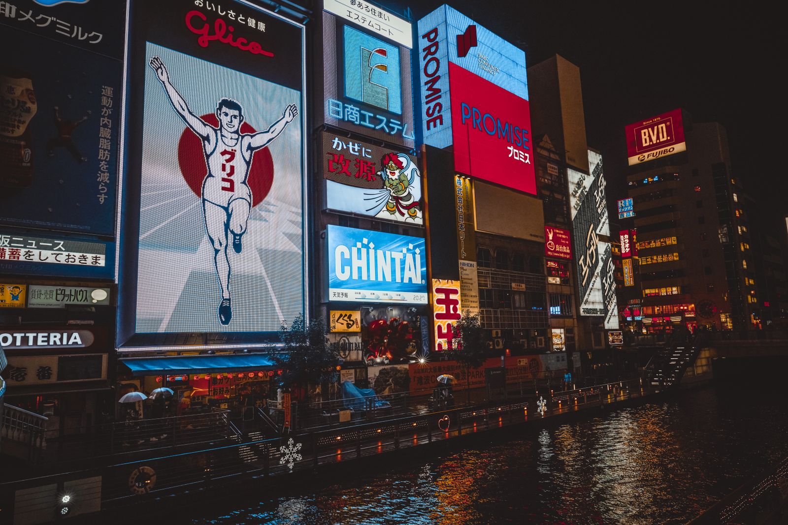 The Dotonbori canal in Osaka at night, the lit Glico running man hoarding on the left and stacked illuminated signs along the far bank reflecting in the water.
