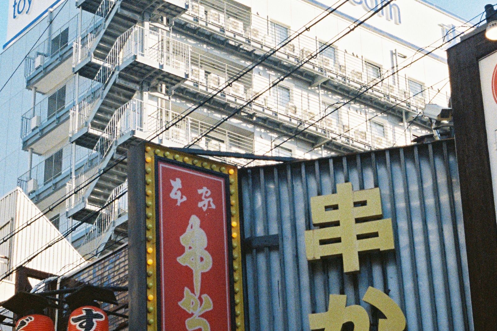 A budget business hotel with an external staircase and stacked balcony rows standing directly above a kushikatsu frontage hung with red lanterns.