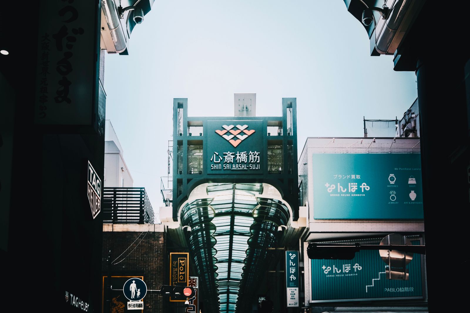 The Shinsaibashi-suji arcade gateway sign with its heart-lattice emblem above the glazed barrel roof of the covered shopping street, seen from the street entrance.