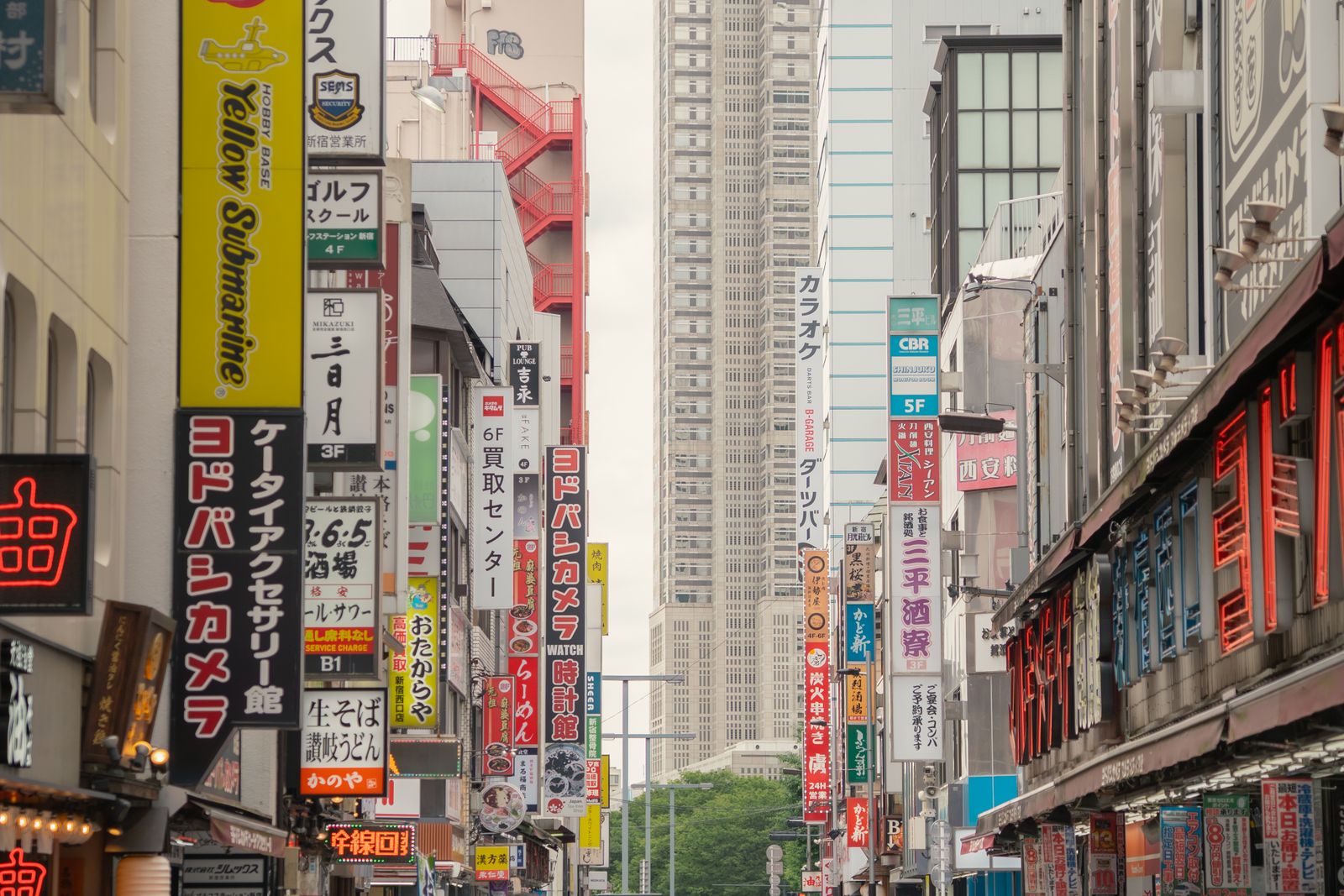 A Shinjuku street at ground level, shopfront signage down both sides and the twin towers of the Tokyo Metropolitan Government Building closing the far end.