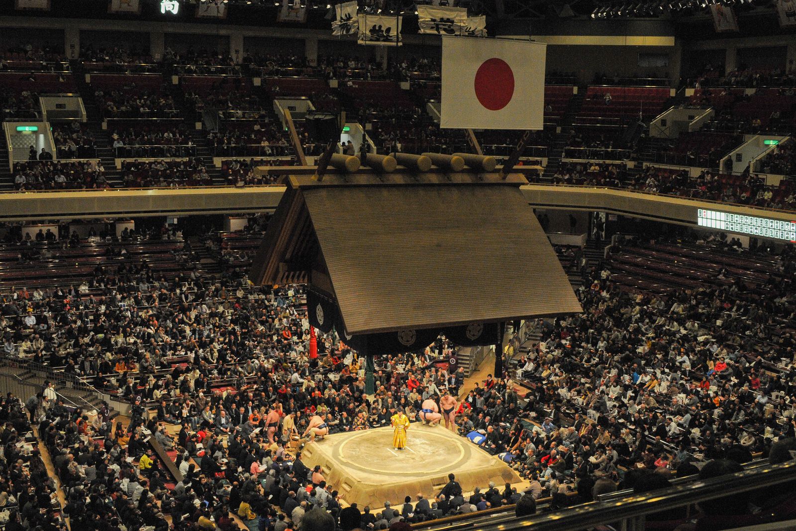 A professional sumo hall at full capacity seen from high in the stands, a suspended tasseled roof over the raised clay ring, the floor laid out as a grid of square four-person box seats with rows of fixed chairs in the galleries above.