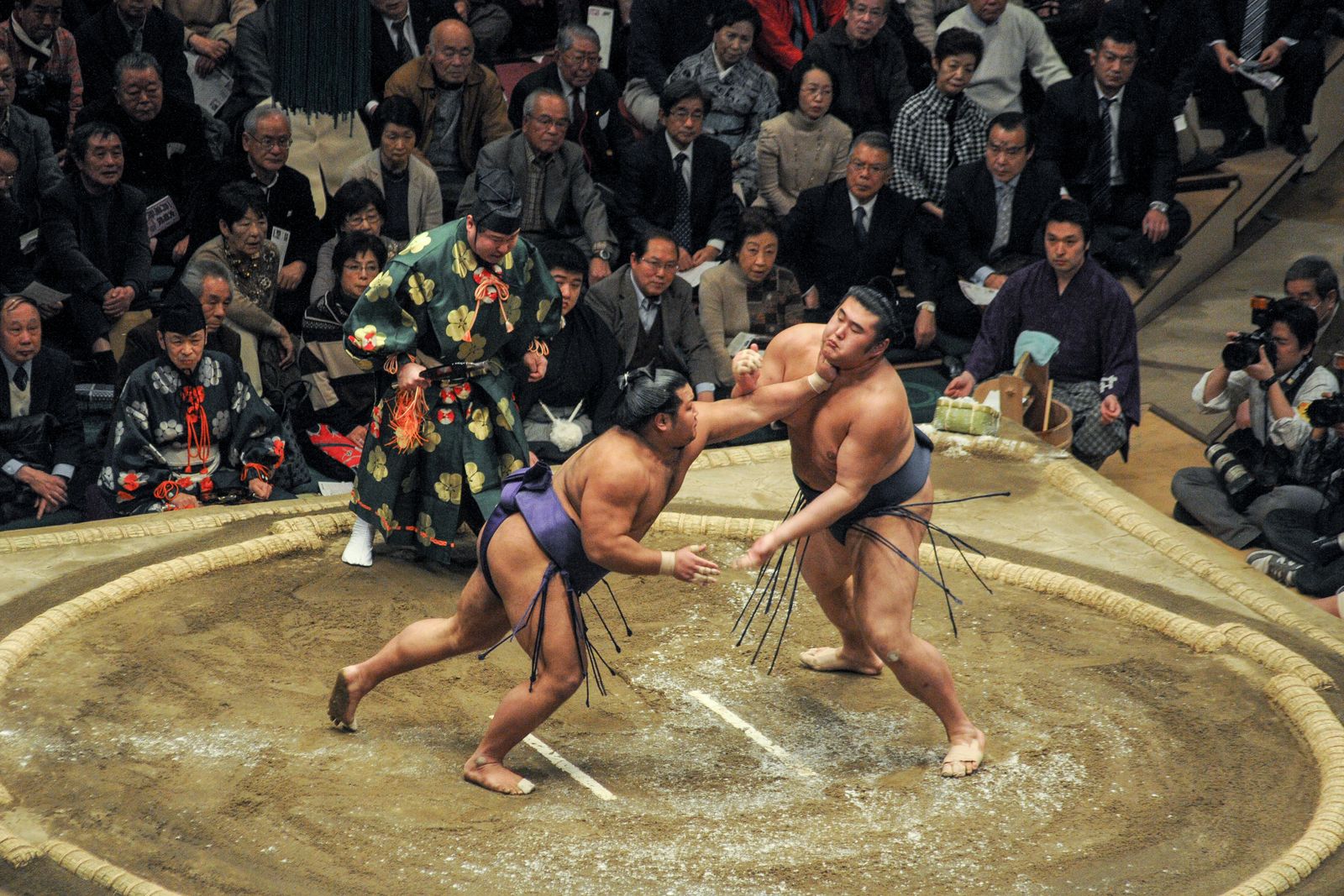Two wrestlers locked at arm's length on the clay, one driving the other back toward the straw bales set into the ring edge, a referee in a green and gold robe behind them.