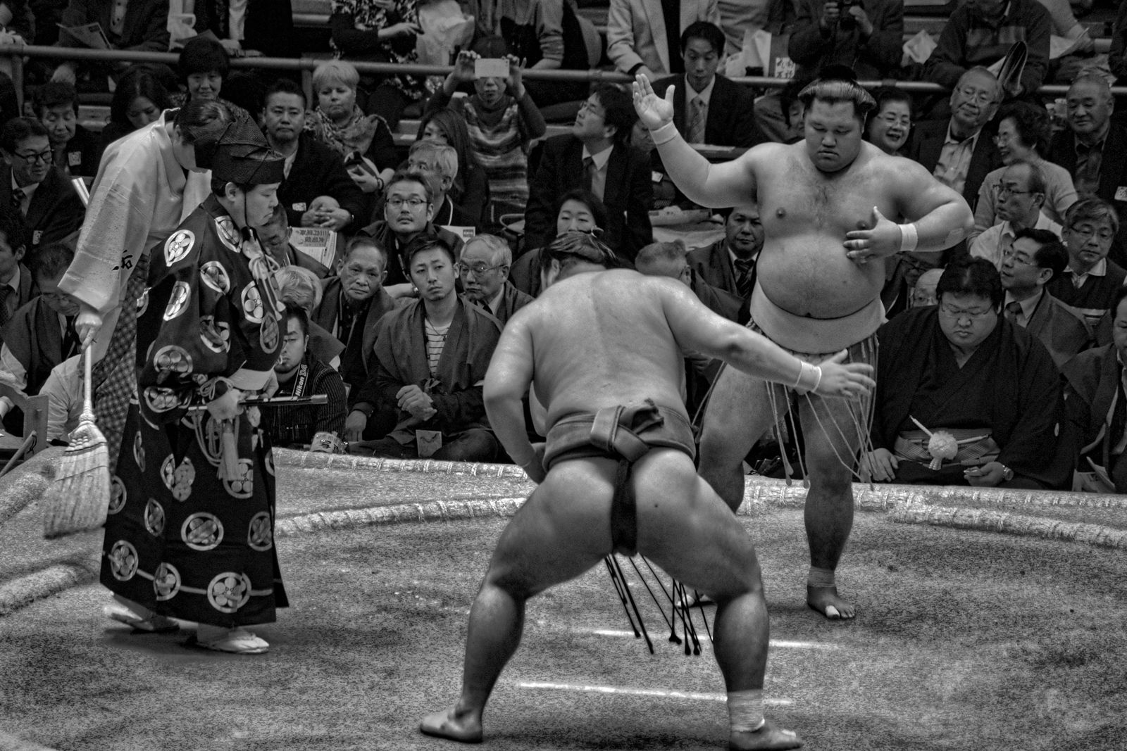 A black and white photograph of a bout at the moment of engagement, one wrestler crouched with his back to the camera, the referee at the left in a full patterned robe holding a war fan.