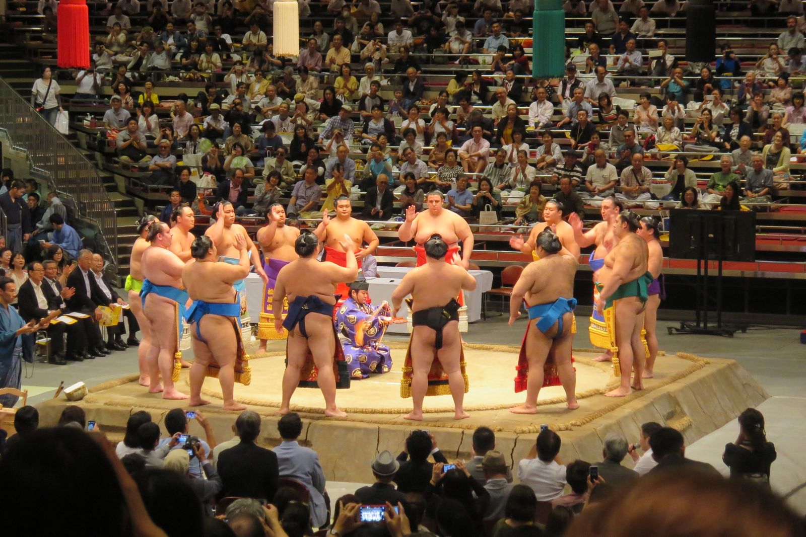 Around a dozen wrestlers in ornate fringed ceremonial aprons standing in a ring around the raised clay dohyo with their arms raised, a referee in a purple robe kneeling at the center rear, a full hall behind.
