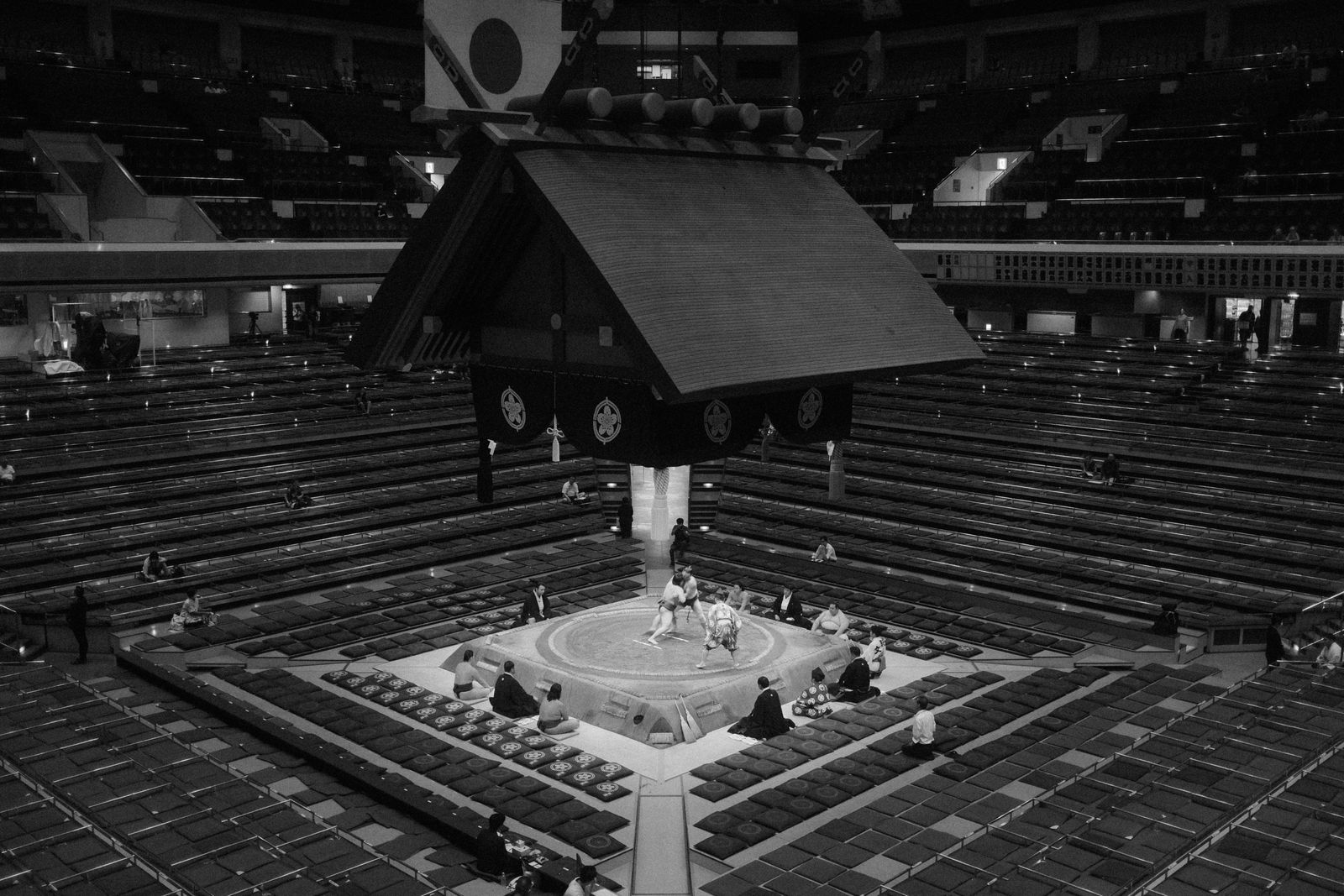A black and white view from high in a sumo hall, a bout under way on the raised ring with a referee beside it, the box-seat floor and the tiered galleries behind almost empty.
