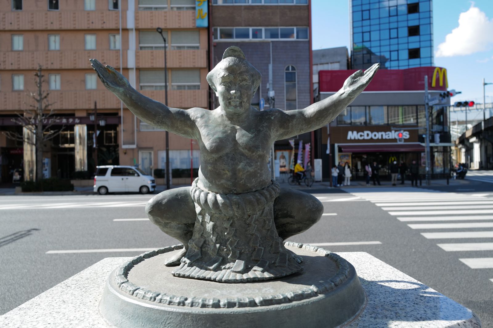 A bronze statue of a sumo wrestler in a low crouch with both arms extended, on a stone plinth cast as a miniature ring, at an ordinary street corner with a crossing, a small van and a traffic signal behind it.