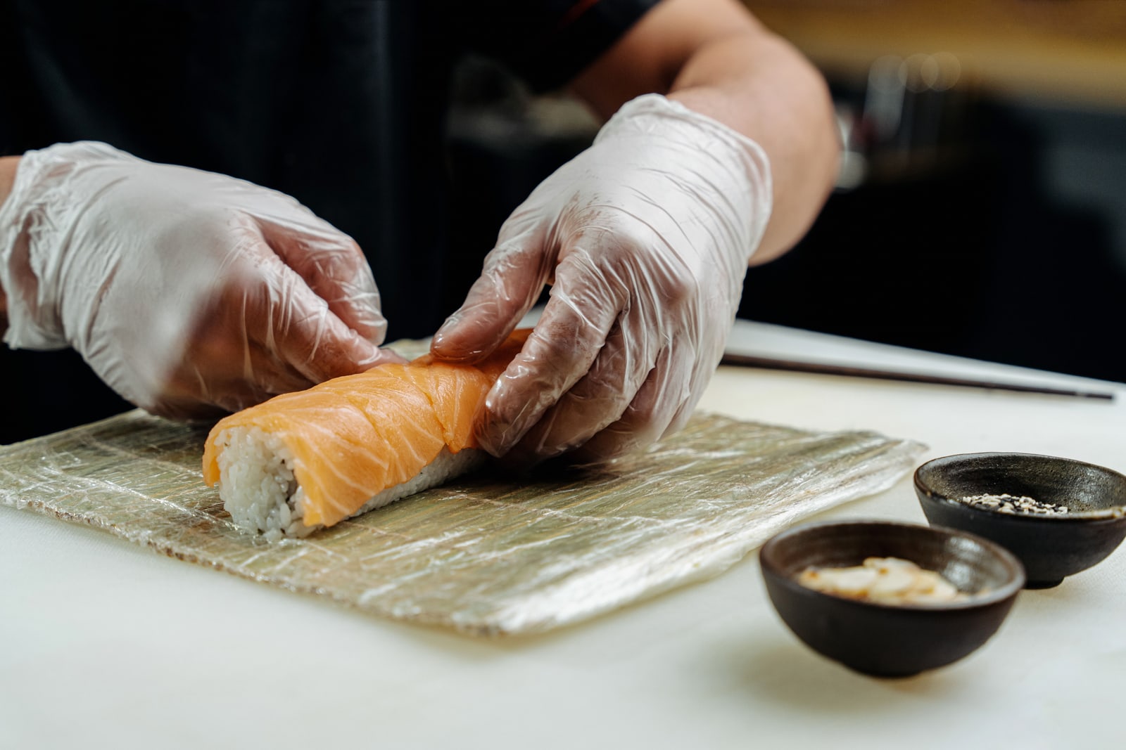 Gloved hands pressing a salmon-topped sushi roll on a bamboo mat, two small dishes of seasoning beside it.