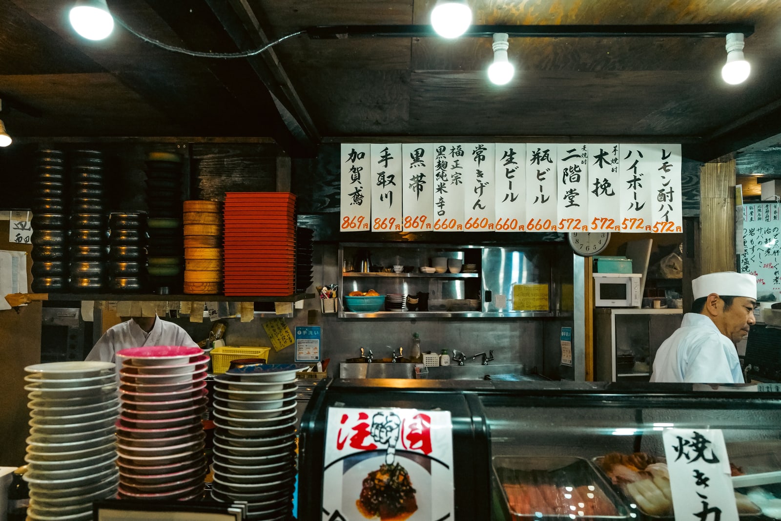 A small sushi bar with stacks of plates, a chef in a white cap behind the counter and handwritten menu strips on the wall above.