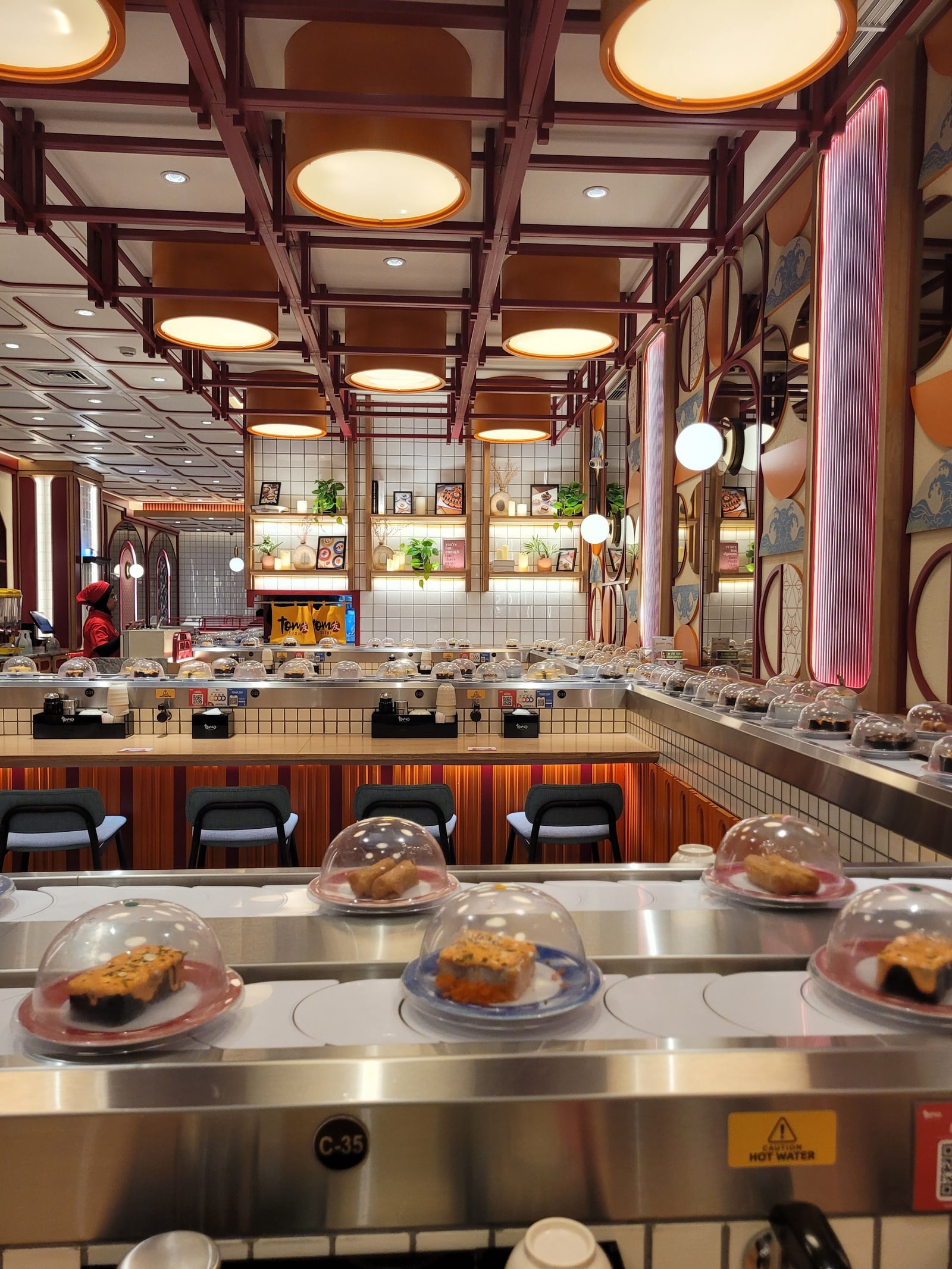 A long conveyor-belt sushi restaurant with plates under clear covers moving along the belt, stools and booths behind it.