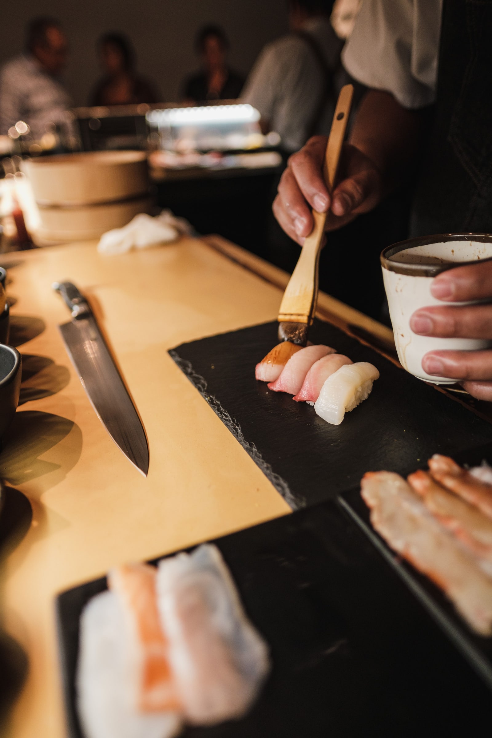 A chef's hands brushing sauce onto nigiri on a black board at a wooden counter, a knife lying beside the board.
