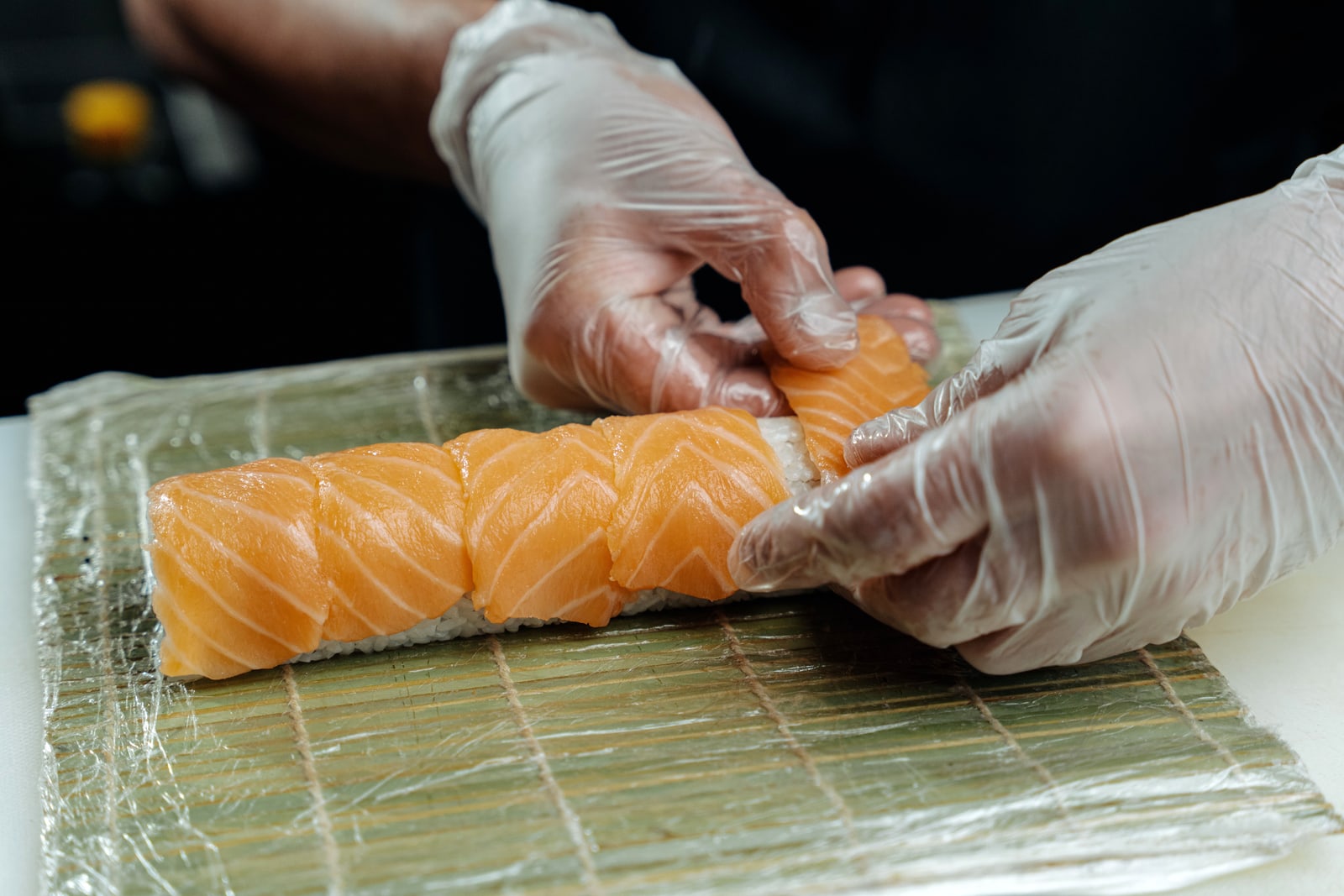 Gloved hands laying slices of salmon over a sushi roll on a bamboo mat.