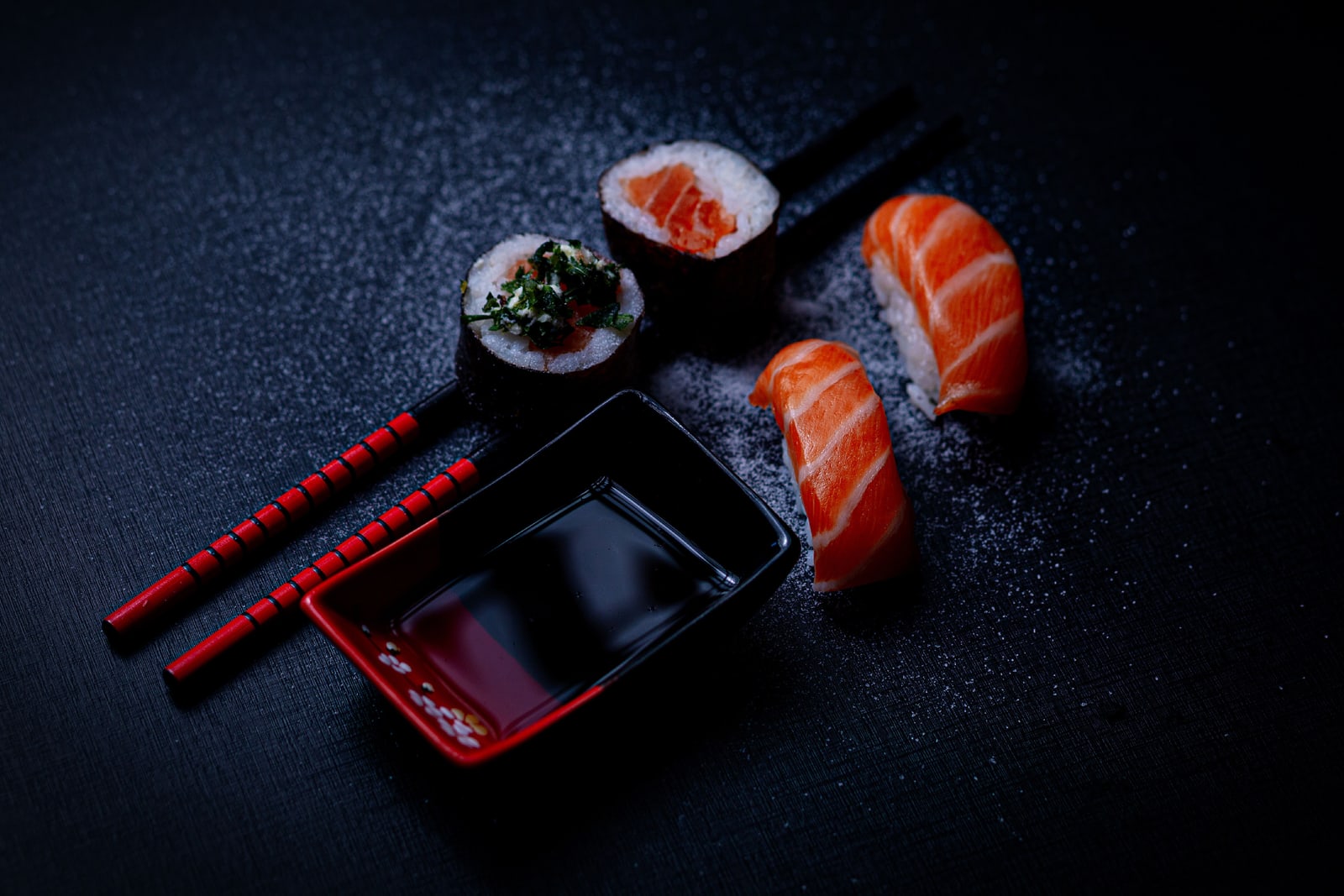 Salmon nigiri and two rolls beside a small dish of soy sauce and a pair of chopsticks on a dark surface.