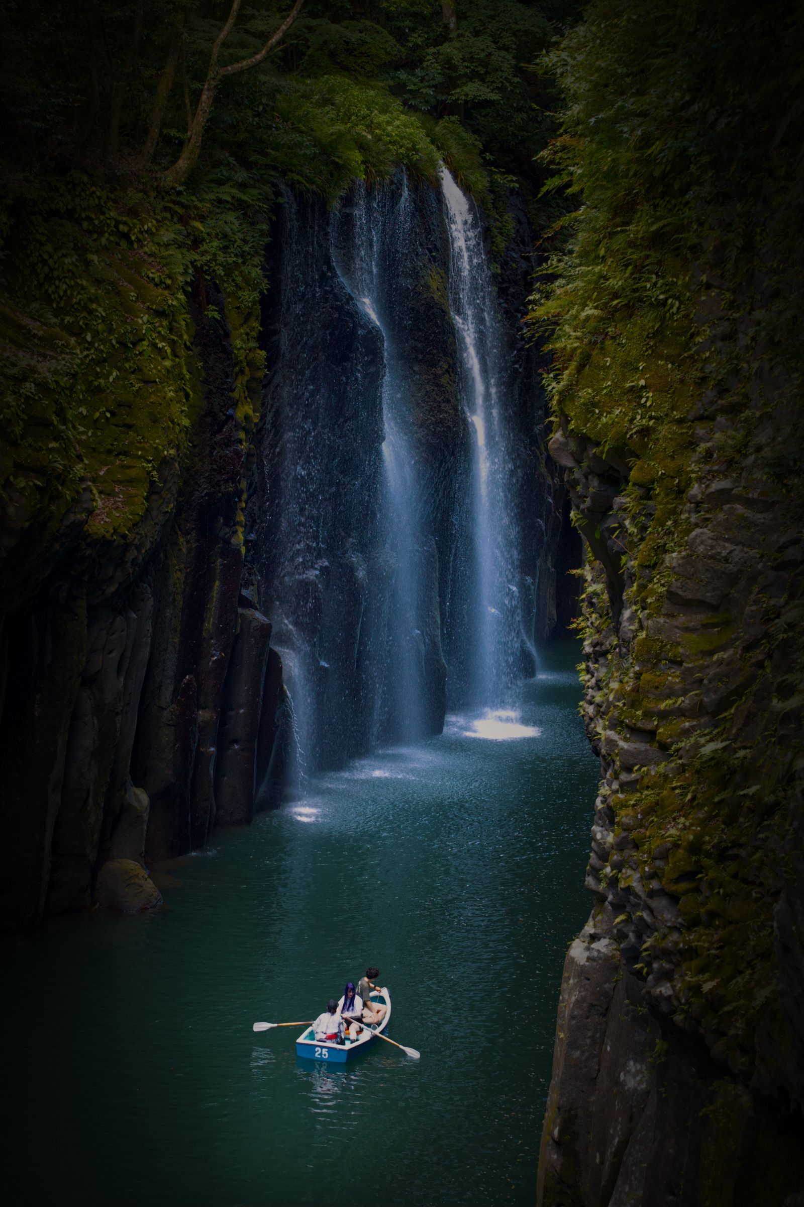A white-and-blue rental rowing boat with three small figures on the green river below Manai Falls in Takachiho Gorge