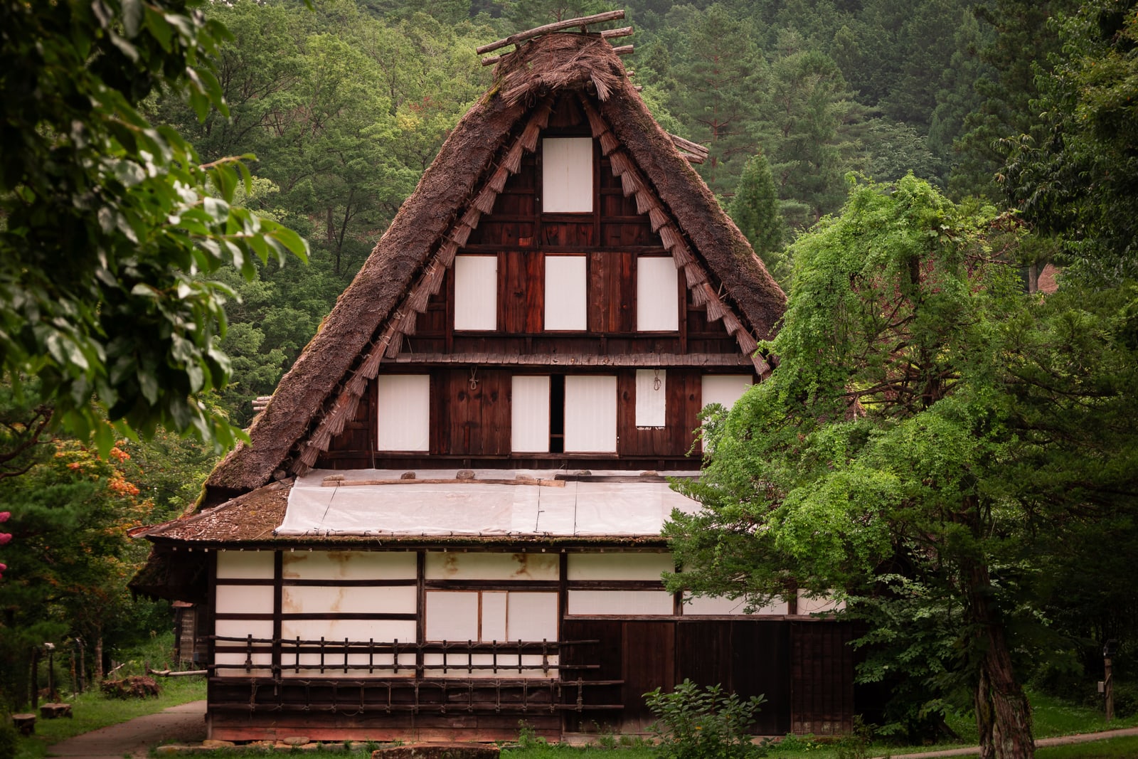 The gable end of a large thatched farmhouse with dark timber and paper-screen windows, green trees and forested hillside around it