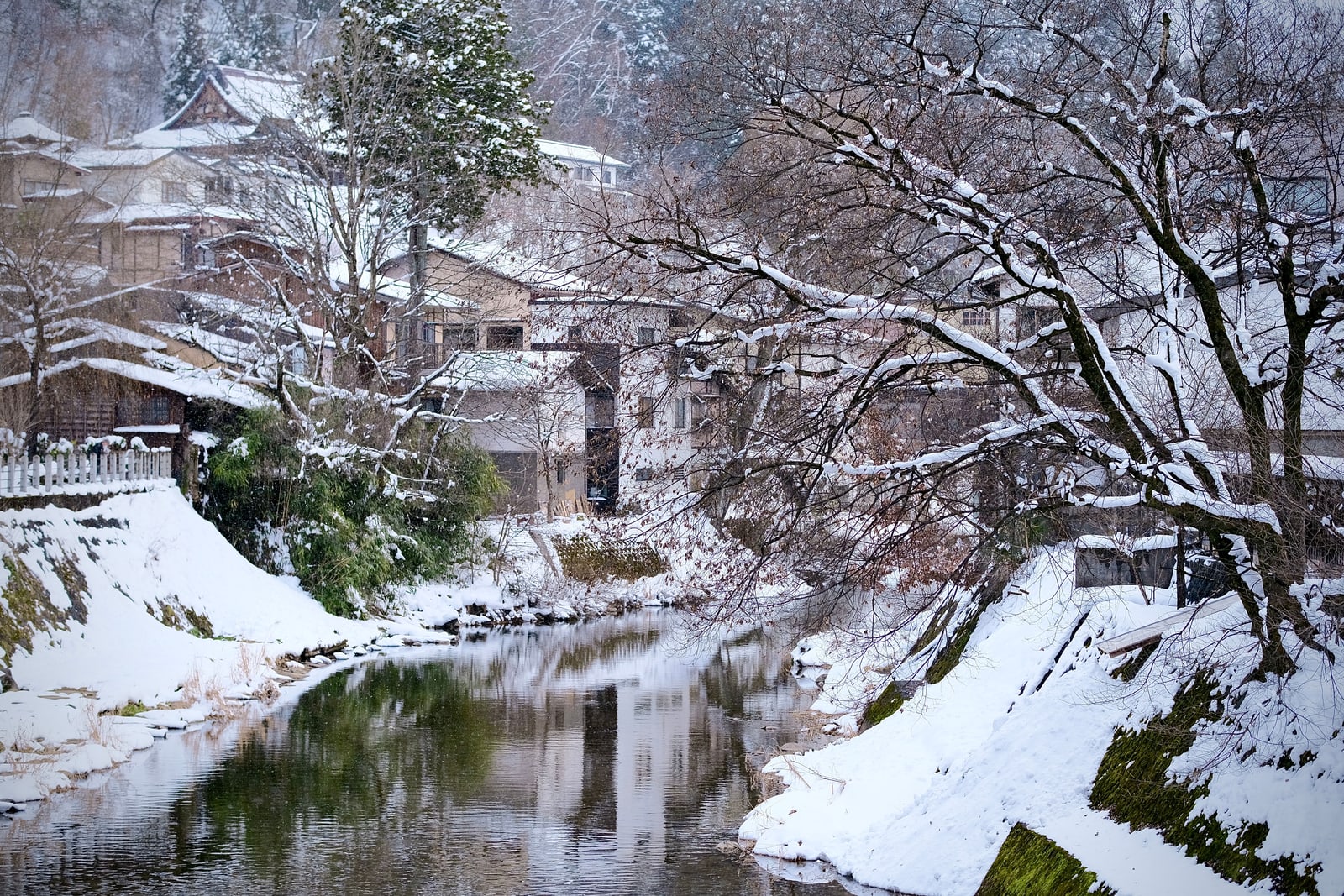 A narrow river between snowy banks in light snowfall, wooden houses with snow-covered roofs and a wooded hillside behind
