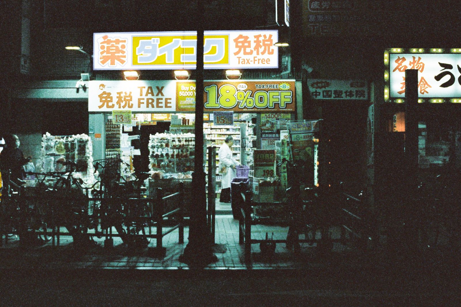 A Japanese drugstore at night, 免税 and TAX FREE lettering across the fascia and a discount panel in the window.