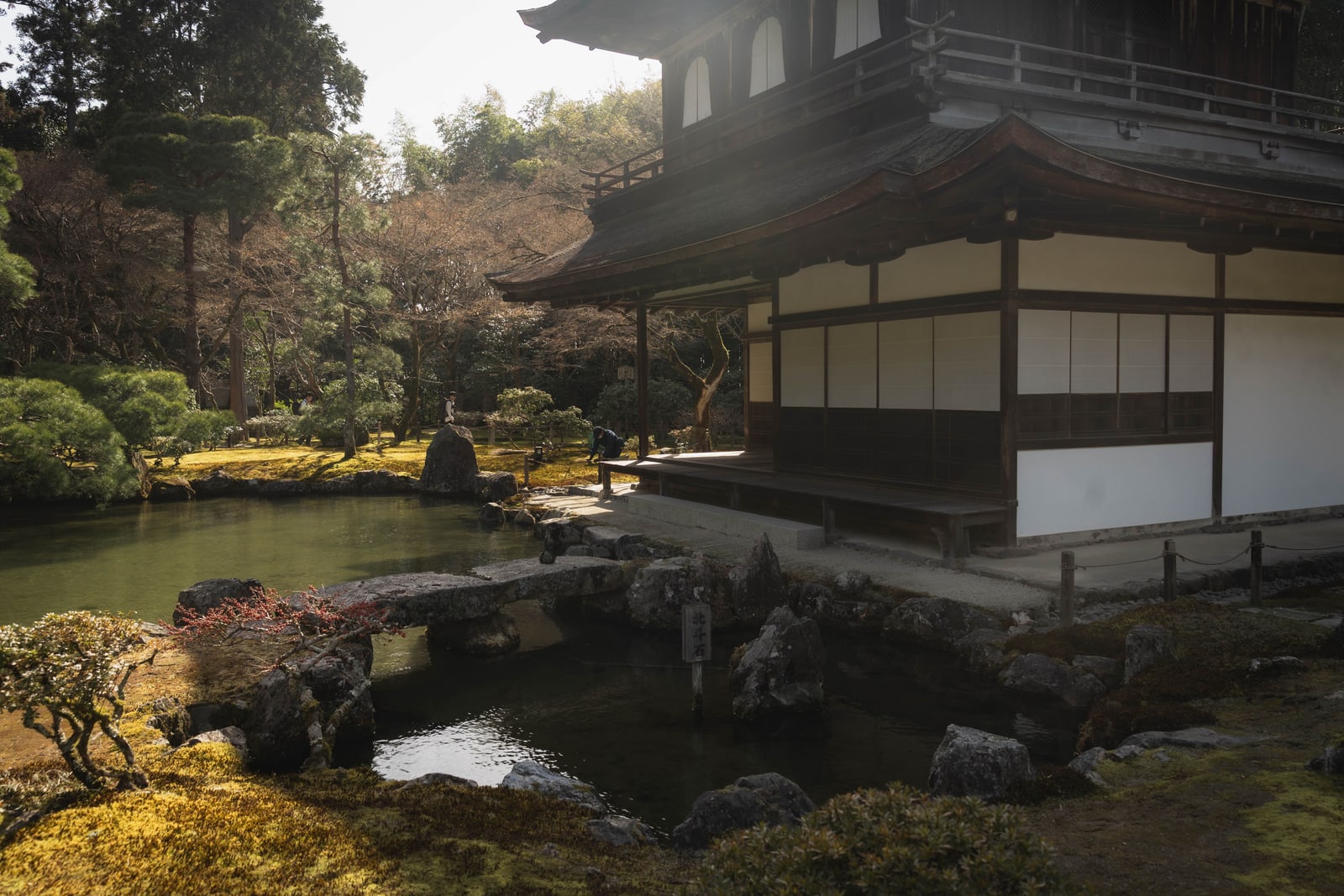 A traditional wooden building beside a pond garden, reached by a stone bridge, in Kyoto.