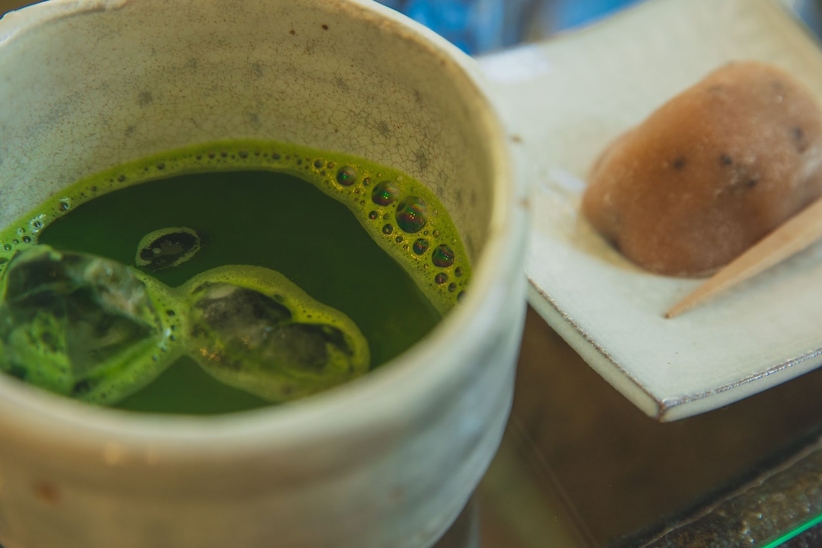 A bowl of whisked matcha beside a wagashi sweet on paper, with a small pick.