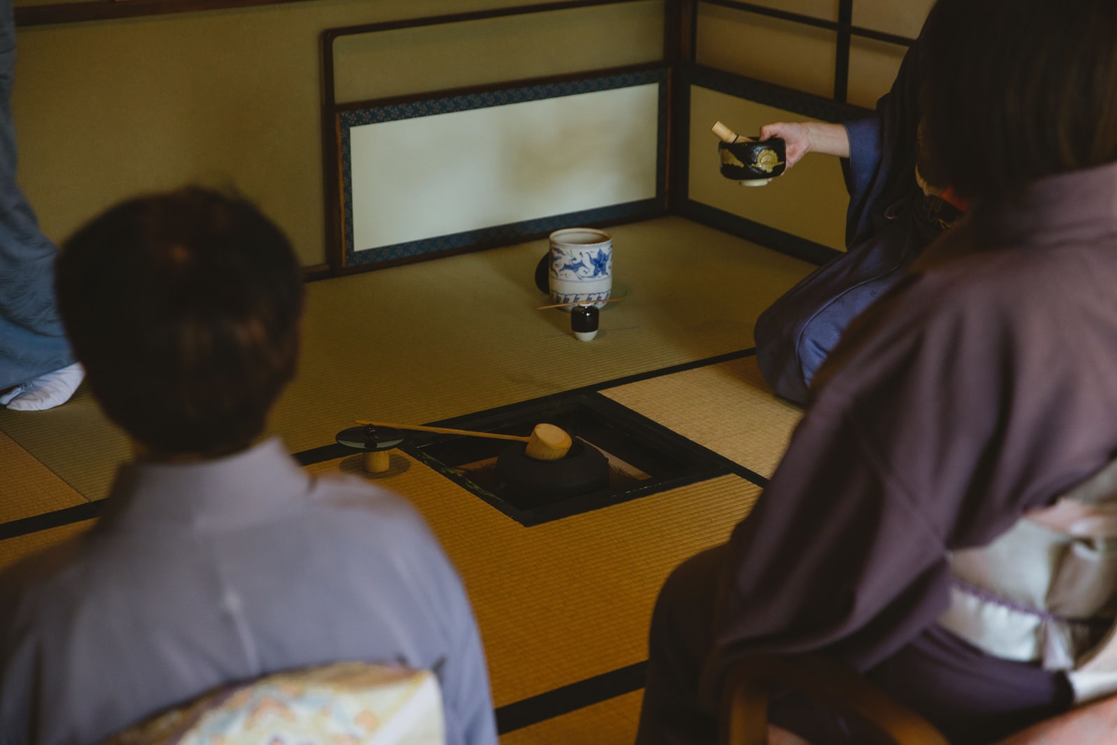 A host in kimono holds a tea bowl out to two guests seated on tatami in a tea room, faces turned away from the camera.