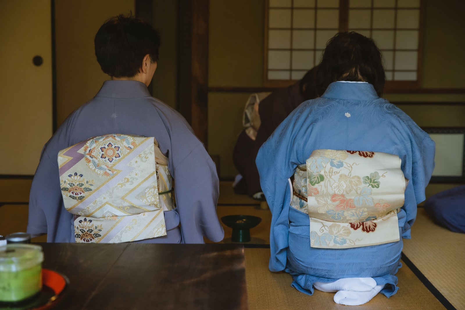 Two people in kimono seen from behind, kneeling on tatami with a tea caddy set before them.