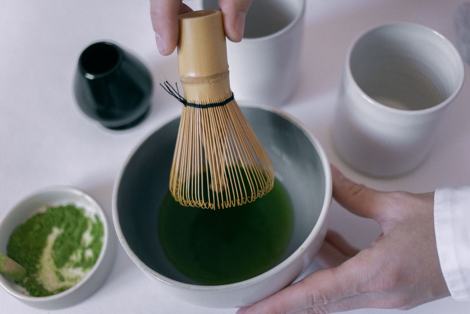 Two hands whisking bright green matcha in a ceramic bowl with a bamboo whisk.