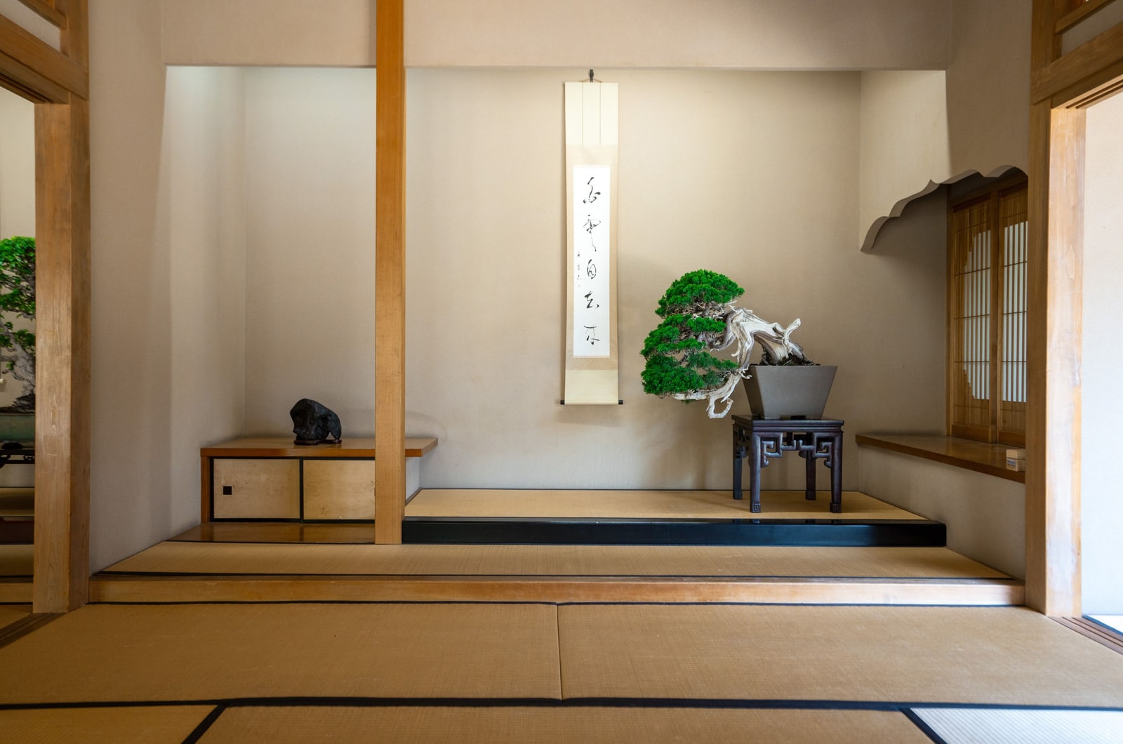 A tokonoma alcove with a hanging calligraphy scroll and a bonsai on a stand, seen past a wooden support post.