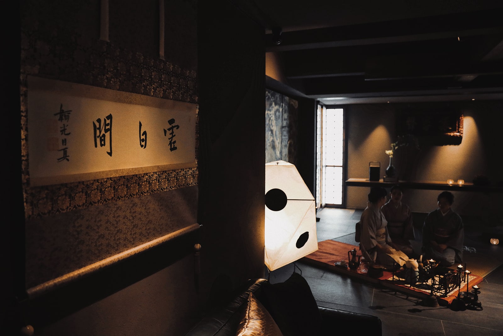 Three people in kimono seated around a low table with tea utensils and candles in a dimly lit room, a hanging scroll on the wall.